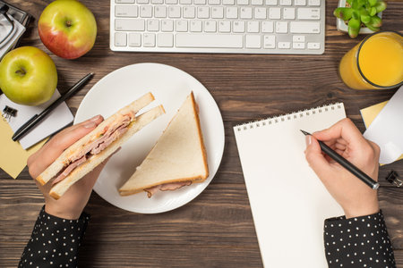 First person top view photo of hands holding sandwich over plate and writing in planner keyboard mouse apples glass of juice plant stationery on isolated dark wooden desk background with blank spaceの写真素材