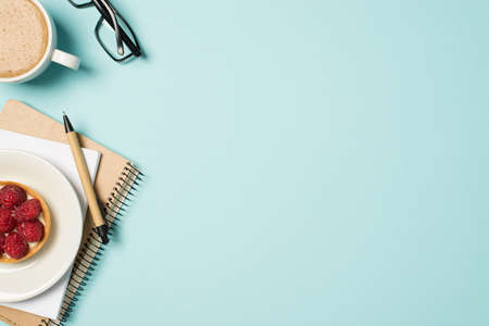 Top view photo of workplace glasses cup of frothy coffee pen copybooks and plate with raspberry cake on isolated pastel blue background with copyspaceの写真素材