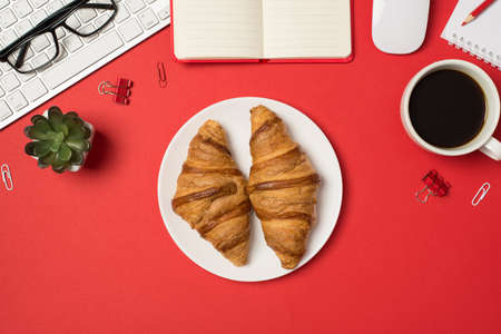 Top view photo of workplace keyboard glasses binder clips flowerpot mouse pencil planners plate with two fresh croissants and cup of coffee on isolated red backgroundの写真素材