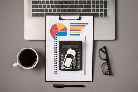 Top view photo of laptop keyboard glasses pen cup of drink reminder clipboard folder with pie chart diagram and car model on calculator on isolated gray backgroundの写真素材