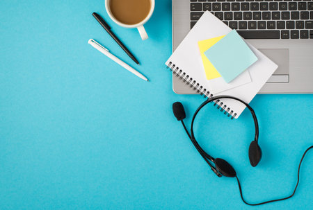 Top view photo of laptop wired headphones with microphone cup of drink pens notepad and sticker notes on isolated pastel blue background with copyspaceの写真素材