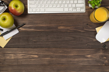Top view photo of workplace white keyboard mouse sticky note paper pen binder clips apples glass of juice and plant on isolated dark wooden table background with copyspace in the middleの写真素材