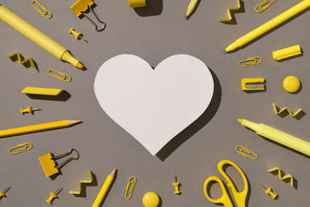 Top view photo of white paper heart surrounded by yellow stationery school supplies felt pens...