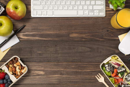Top view photo of workplace two lunchboxes with healthy food fork stationery keyboard mouse apples glass of juice plant in the corners isolated dark wooden desk background with copyspace in the middleの写真素材