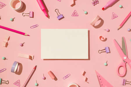 Overhead photo of scissors tape paper clips triangle pins spirals pencil pen eraser marker stickers and white card in the middle isolated on the pink backdropの写真素材