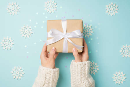 First person top view photo of hands in white sweater holding craft paper giftbox with white ribbon bow over big decorative snowflakes and shiny confetti on isolated pastel blue backgroundの写真素材