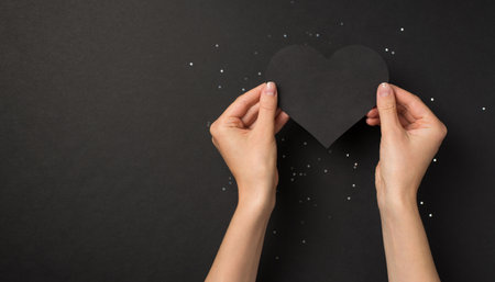 First person top view photo of woman hands holding black paper heart over sequins on isolated black background with copyspaceの写真素材