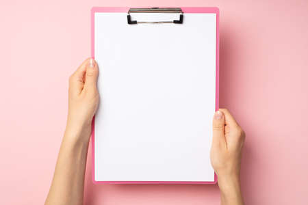 First person top view photo of hands holding pink clipboard with paper sheet on isolated pastel pink background with blank spaceの写真素材