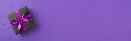 Top view photo of stylish black giftbox with purple ribbon bow and confetti on isolated violet background with copyspaceの写真素材