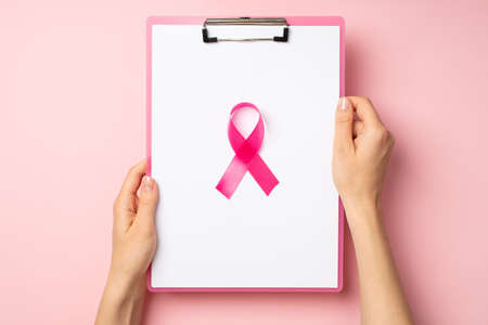 First person top view photo of woman's hands holding pink clipboard with paper sheet and pink ribbon symbol of breast cancer awareness in the middle on isolated pastel pink backgroundの写真素材
