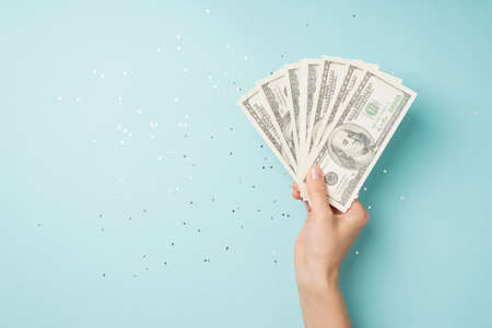 First person top view photo of hand holding fan of money dollars over shiny sequins on isolated pastel blue background with copyspaceの写真素材