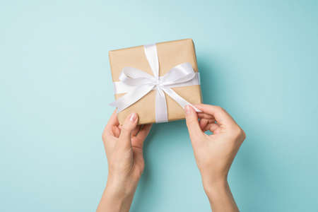 First person top view photo of hands unpacking craft paper giftbox with white ribbon bow on isolated pastel blue backgroundの写真素材