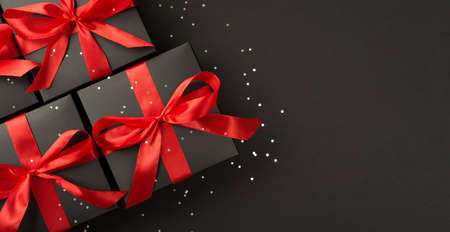 Top view photo of black gift boxes with red ribbon bow and sequins on isolated black background with blank spaceの写真素材