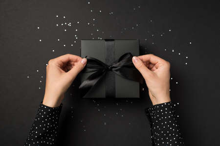First person top view photo of female hands in polka dot shirt tying black giftbox with black ribbon bow over shiny confetti on isolated black backgroundの写真素材