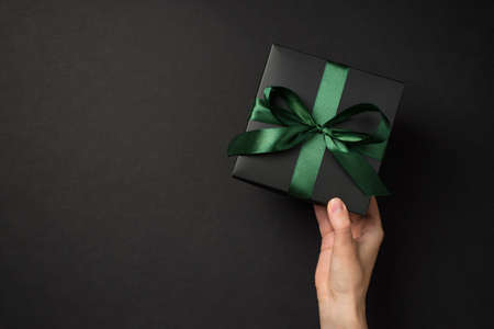 First person top view photo of hand holding black giftbox with green satin ribbon bow on isolated black background with copyspaceの写真素材