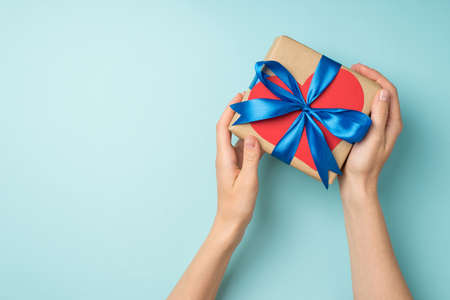 First person top view photo of female hands holding craft paper giftbox with blue ribbon bow and red paper heart on isolated pastel blue background with copyspaceの写真素材