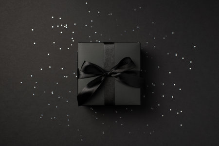 Top view photo of black giftbox with black satin ribbon bow over shiny sequins on isolated black backgroundの写真素材