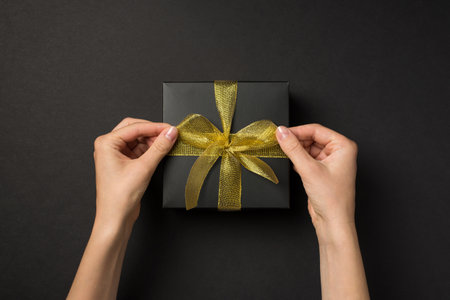 First person top view photo of girl's hands tying golden mesh ribbon bow on black gift box on isolated black backgroundの写真素材