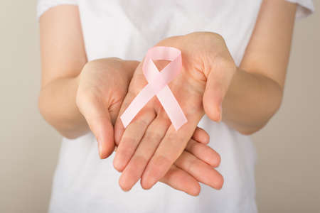 Photo of girl in white t-shirt holding pastel pink ribbon in palms symbol of breast cancer awareness on isolated grey backgroundの写真素材