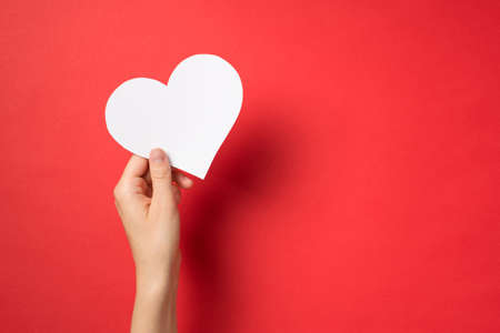 First person top view photo of hand holding white paper heart on isolated red background with...