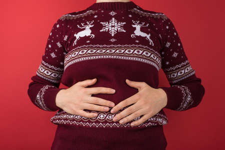 Cropped closeup photo of girl in red and white christmas sweater holding her hurting stomach on isolated red backgroundの写真素材