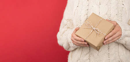 Cropped closeup photo of girl in white knitted jumper holding craft paper giftbox with twine bow on...