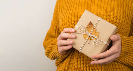 Photo of woman in yellow knitted sweater holding craft paper giftbox with twine bow and orange autumn leaf on isolated white backgroundの写真素材