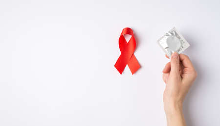 First person top view photo of female hand holding silver condom pack over red ribbon symbol of aids...