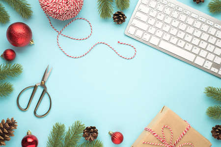 Top view photo of keyboard, pine twigs cones, red christmas tree balls, scissors, spool of decorative rope and craft paper giftbox on isolated pastel blue background with copyspace in the middleの写真素材