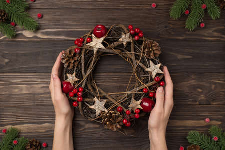 First person top view photo of pine branches and female hands holding christmas twig wreath with...