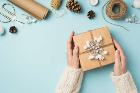 First person top view photo of woman's hands in knitted pullover holding giftbox decorated with snow...