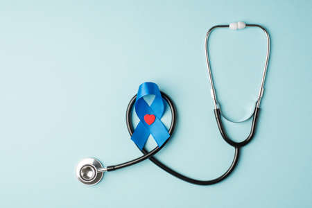 Top view photo of stethoscope and blue silk ribbon with small red heart symbol of prostate cancer awareness on isolated pastel blue background with blank spaceの写真素材