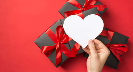 First person top view photo of hand holding white paper heart over stack of gift boxes on isolated...