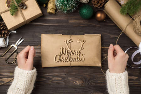 First person top view photo of woman's hands in sweater decorating kraft paper package with merry christmas wooden text and twine balls pine twigs handicraft tools on isolated wooden desk backgroundの写真素材