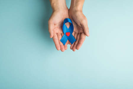 First person top view photo of male hands demonstrating blue ribbon with small red heart in palms symbol of prostate cancer awareness on isolated pastel blue background with empty spaceの写真素材