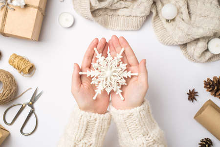 First person top view photo of female hands in sweater holding big decorative snowflake giftbox scarf christmas tree balls cones anise candles scissors spools of twine on isolated white backgroundの写真素材