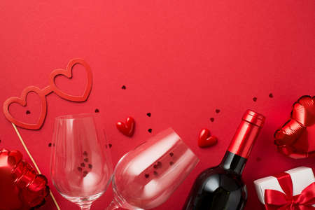 Top view photo of valentine's day decorations gift box heart shaped paper glasses balloons two wineglasses wine bottle and red confetti on isolated red background with copyspaceの写真素材