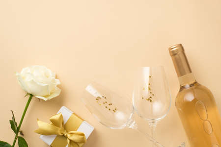 Top view photo of white rose two wineglasses with golden sequins bottle of white wine and white giftbox with gold ribbon bow on isolated beige background with empty spaceの写真素材