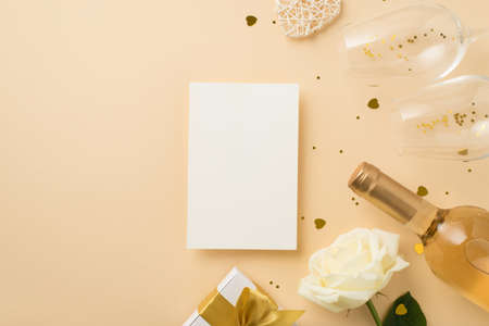 Top view photo of paper sheet white giftbox with gold bow two wineglasses wine bottle sequins white rose and heart shaped golden confetti on isolated beige background with blank spaceの写真素材
