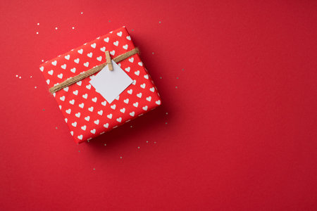 Top view photo of valentine's day decorations shiny sequins giftbox in red wrapping paper with heart pattern and twine with pinned note on isolated red background with blank spaceの写真素材