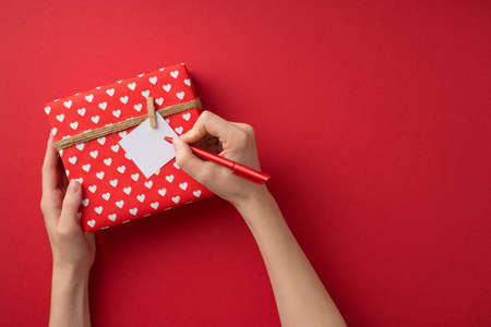 First person top view photo of valentine's day decor female hands writing love letter attached to giftbox in red wrapping paper with heart pattern and twine on isolated red background with empty spaceの写真素材