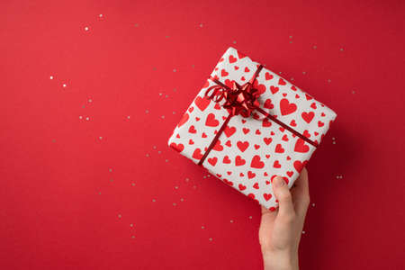 First person top view photo of st valentine's day decor female hand giving giftbox in white wrapping paper with heart pattern red star bow and sequins on isolated red background with empty spaceの写真素材