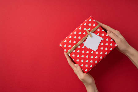 First person top view photo of valentine's day decorations woman's hands giving giftbox in red wrapping paper with heart pattern twine and pinned letter on isolated red background with empty spaceの写真素材