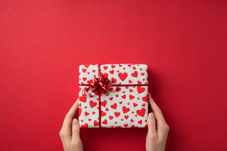 First person top view photo of valentine's day decorations young person's hands holding giftbox in white wrapping paper with heart pattern and red star bow on isolated red background with empty spaceの写真素材