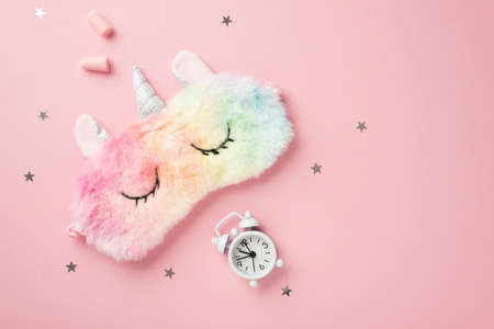 Top view photo of funny multicolored fluffy unicorn sleeping mask small white alarm clock earplugs and star shaped confetti on isolated pastel pink background with copyspaceの写真素材
