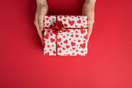 First person top view photo of st valentine's day decorations woman's hands giving gift box in white wrapping paper with heart pattern and red star bow on isolated red background with empty spaceの写真素材