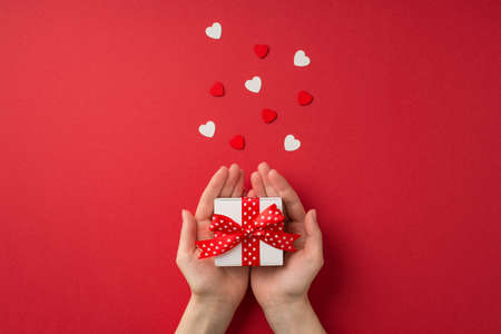 First person top view photo of valentine's day decorations female hands holding small white giftbox with red dotted ribbon bow on palms and hearts on isolated red background with copyspaceの写真素材