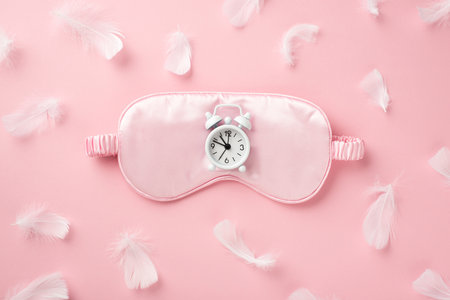 Top view photo of small white alarm clock on pink silk sleeping mask and light pink feathers on isolated pastel pink backgroundの写真素材