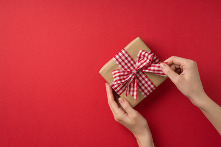 First person top view photo of valentine's day decorations woman's hands untying checkered ribbon bow on craft paper giftbox on isolated red background with copyspaceの写真素材