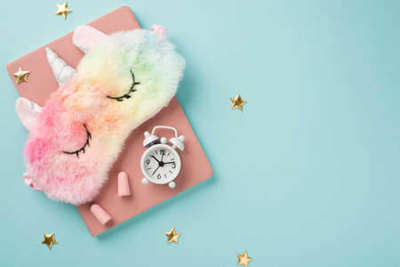 Top view photo of cute colorful fluffy eye mask in shape of unicorn on pink copybook with white alarm clock and pink ear plugs on pastel blue isolated background with golden stars and copyspaceの写真素材